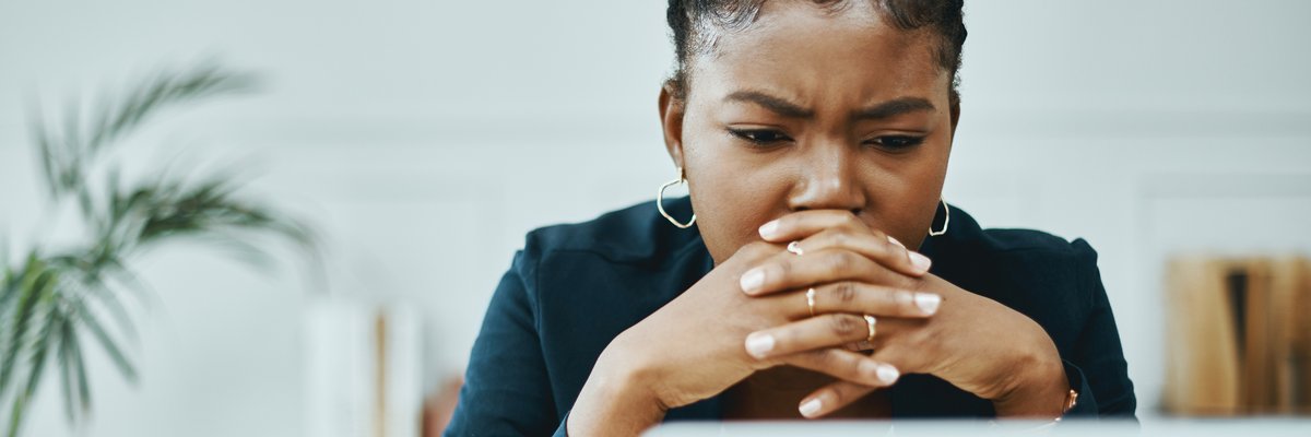 A young business professional worriedly looks at a computer screen as their head rests on their clasped hands.