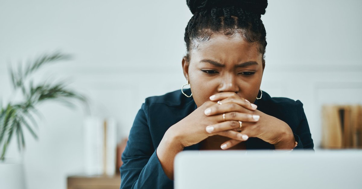 A young business professional worriedly looks at a computer screen as their head rests on their clasped hands.