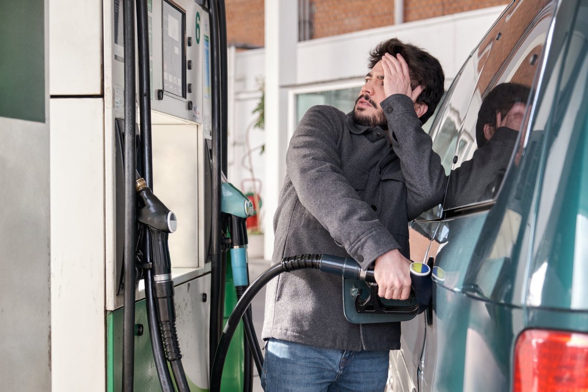 A young adult refuels their car while looking worried at the high gas prices at a gas station.