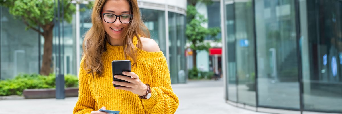 Person smiling while holding credit card and cellphone outdoors near street.