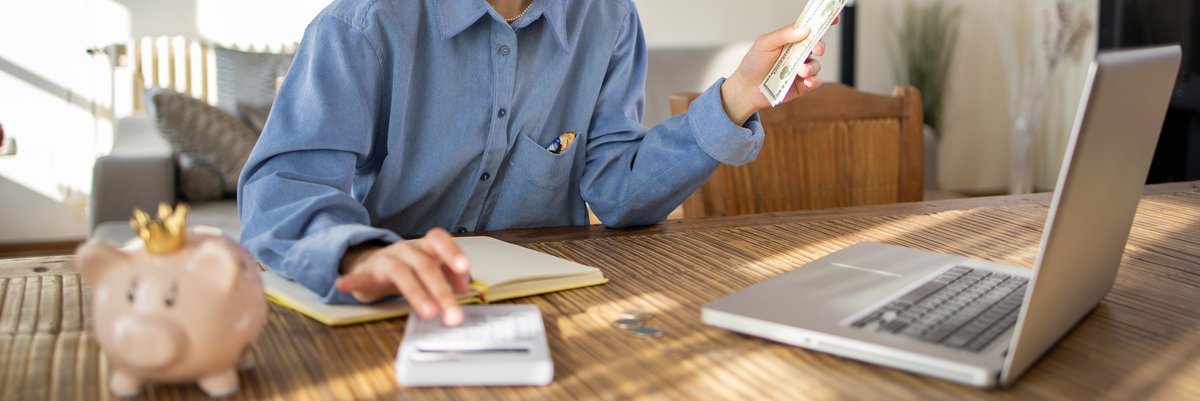 Person holding money with laptop, notebook, and piggy bank nearby.