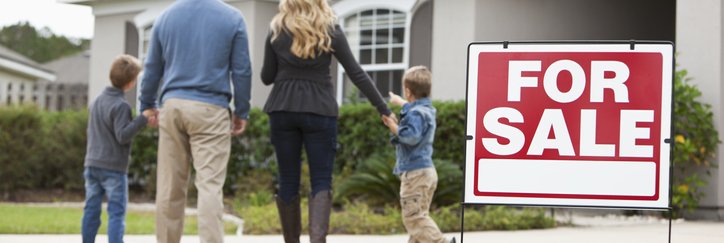 A family looks excitedly at a home with a For Sale sign on the lawn.