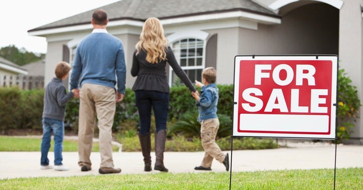 A family looks excitedly at a home with a For Sale sign on the lawn.