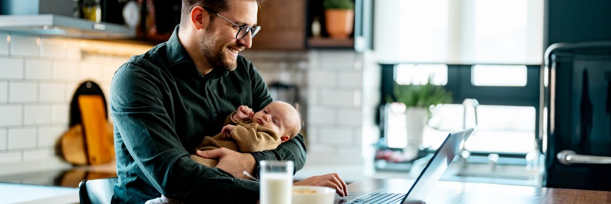 Man holding infant while using laptop and having a bowl of cereal and glass of milk.