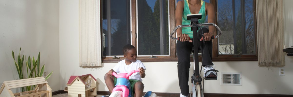 A parent uses an exercise bike at home while their young child sits on their own tricycle.