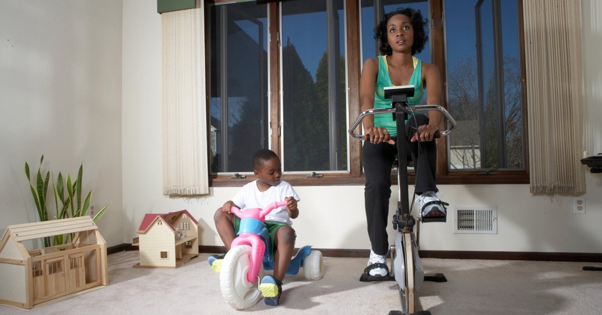 A parent uses an exercise bike at home while their young child sits on their own tricycle.