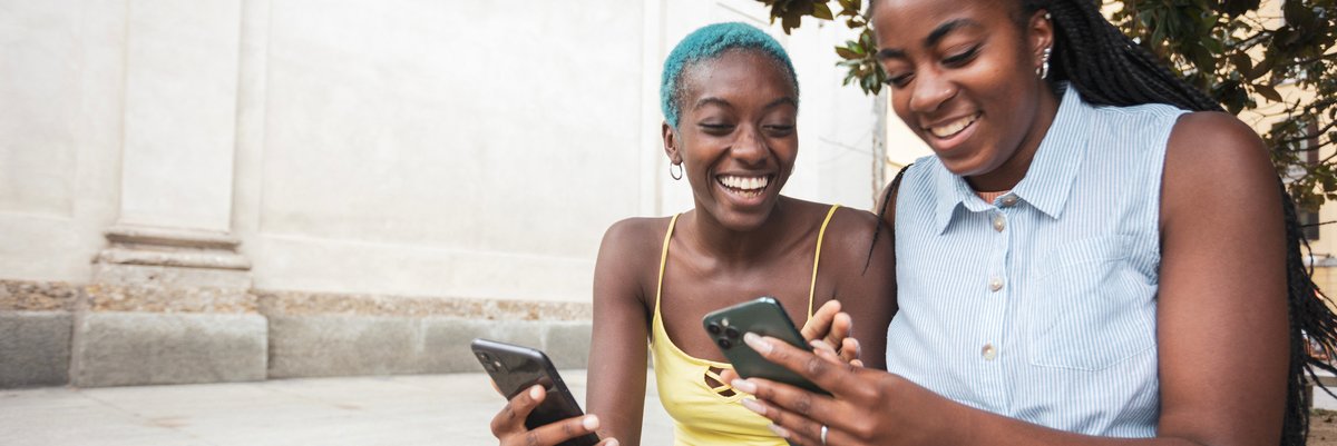 Two women outdoors smiling looking at their cellphones.