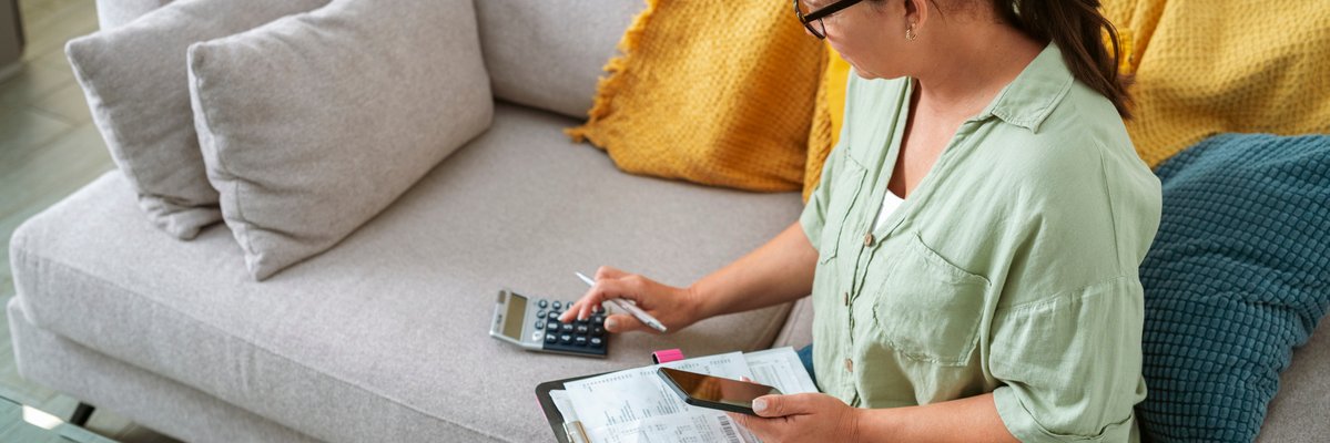 Person holding phone and paperwork on a clipboard and making calculations while sitting on a couch.