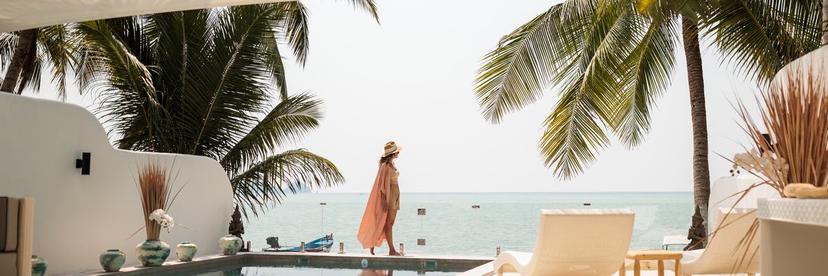 Woman in resort wear walking by a pool with ocean in background.