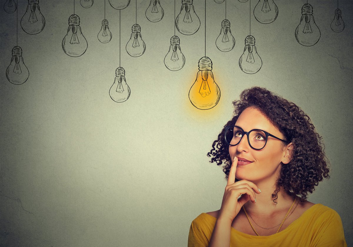Woman in glasses looking up at drawings of many light bulbs, only one of which is lit up.