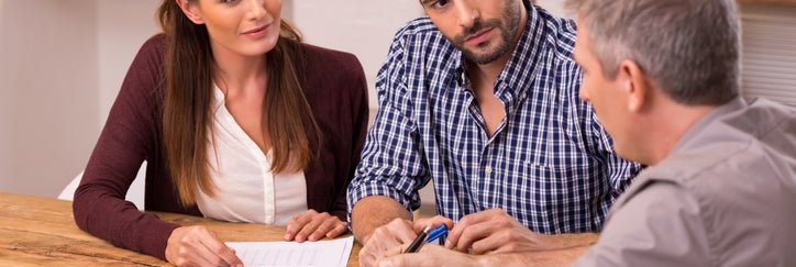 Couple sitting at wooden table discussing paperwork with advisor.