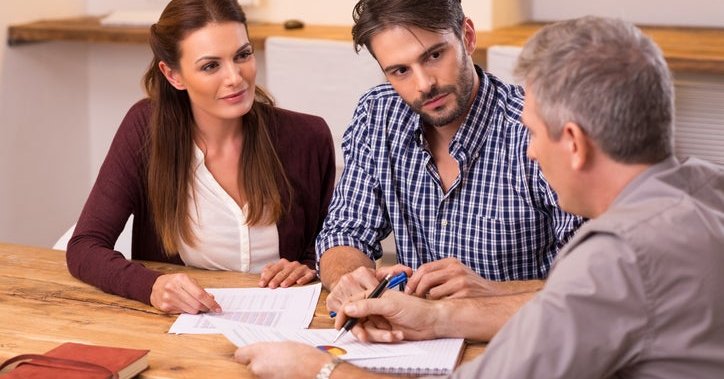 Couple sitting at wooden table discussing paperwork with advisor.