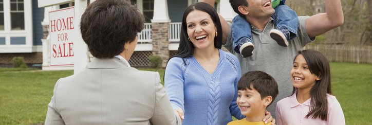 A parent shakes hands with a realtor as their family celebrates. A house with a sign that reads "Home for Sale" is behind them.