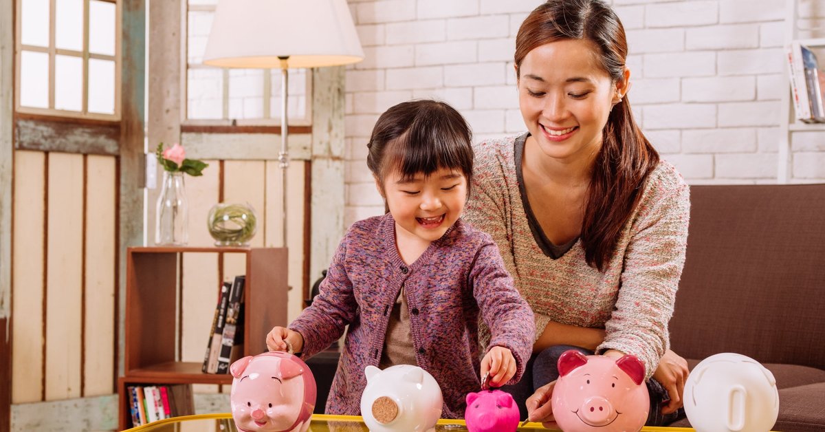 A child puts coins into differently sized piggy banks under the happy supervision of her parent.