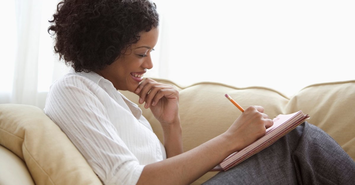 A person reclines on their couch while writing with a notepad.
