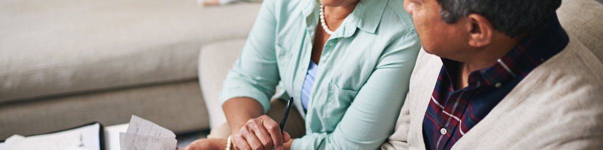 An older couple uses a laptop, calculator, and papers to budget while sitting on the living room sofa