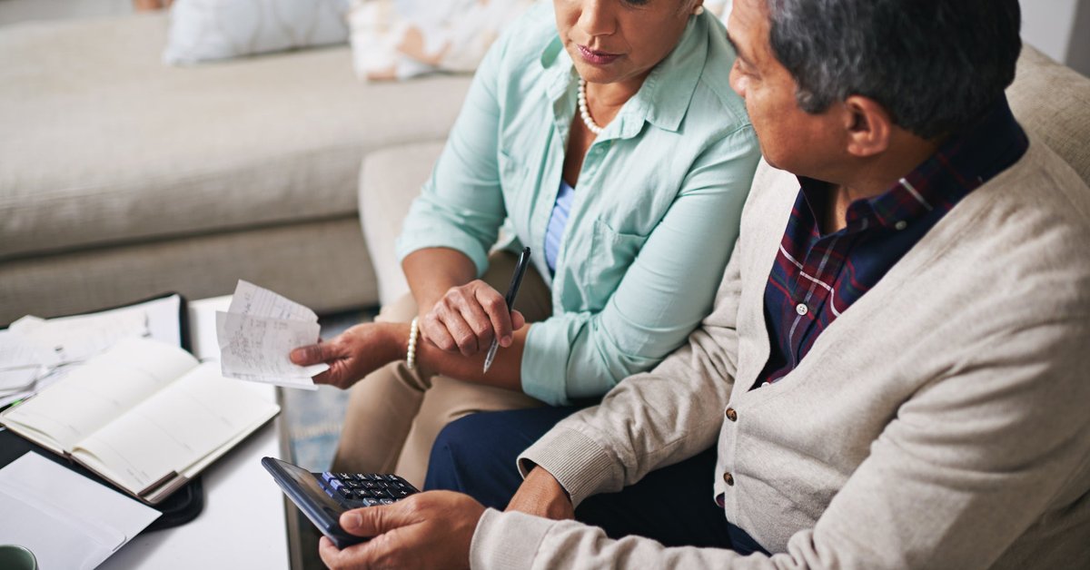 An older couple uses a laptop, calculator, and papers to budget while sitting on the living room sofa