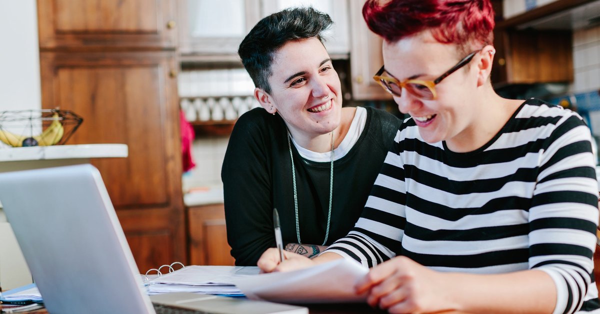 A couple smiles as they pay bills online at the kitchen table.