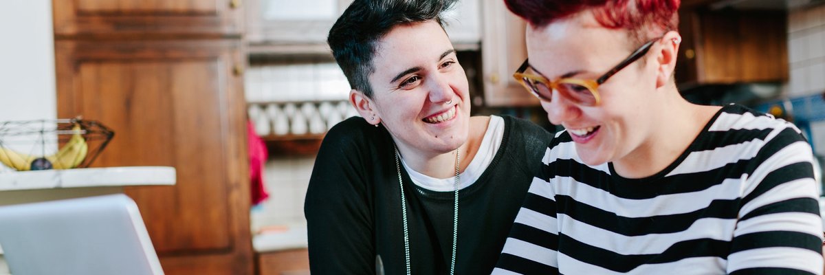 A couple smiles as they pay bills online at the kitchen table.