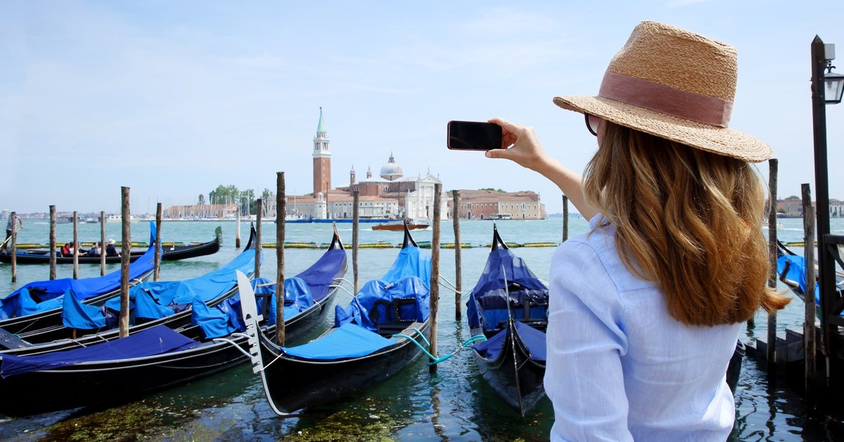 Woman in hat facing away and taking photos of an enormous old church across a bay.
