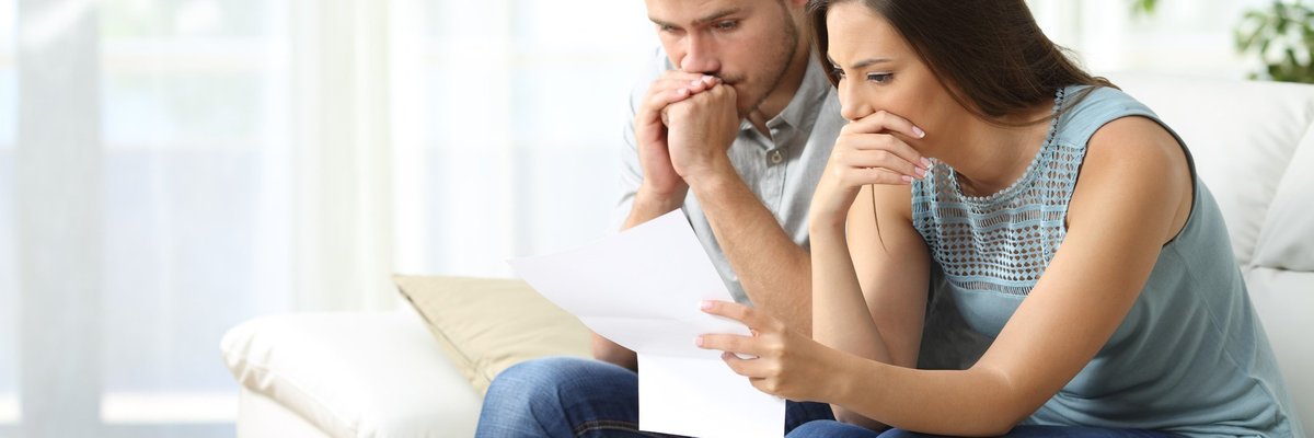 Man and woman sitting on couch looking worried with paperwork in hand