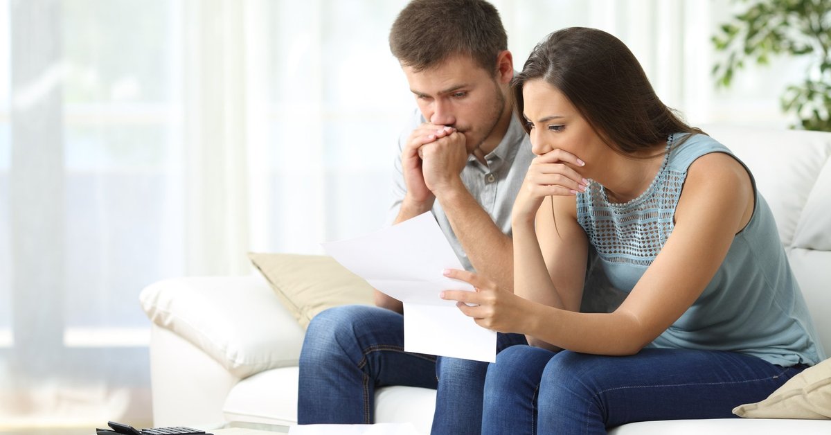 Man and woman sitting on couch looking worried with paperwork in hand