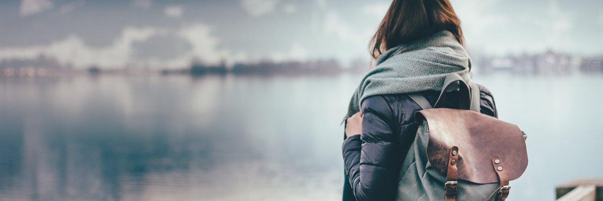 Intrepid woman wearing backpack facing away toward mountain lake.