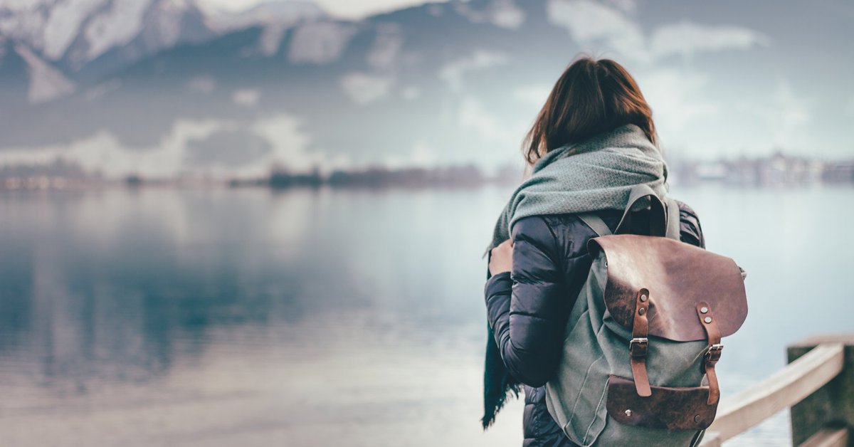 Intrepid woman wearing backpack facing away toward mountain lake.