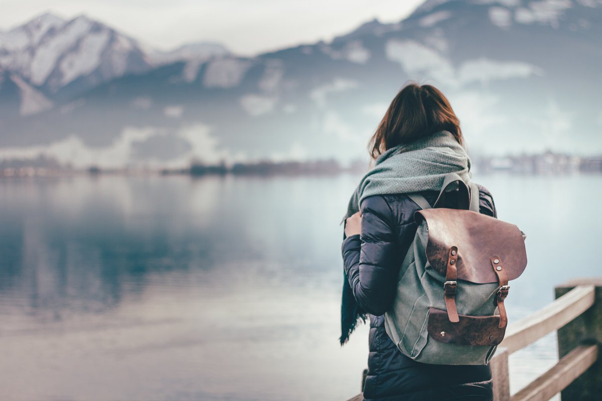 Intrepid woman wearing backpack facing away toward mountain lake.