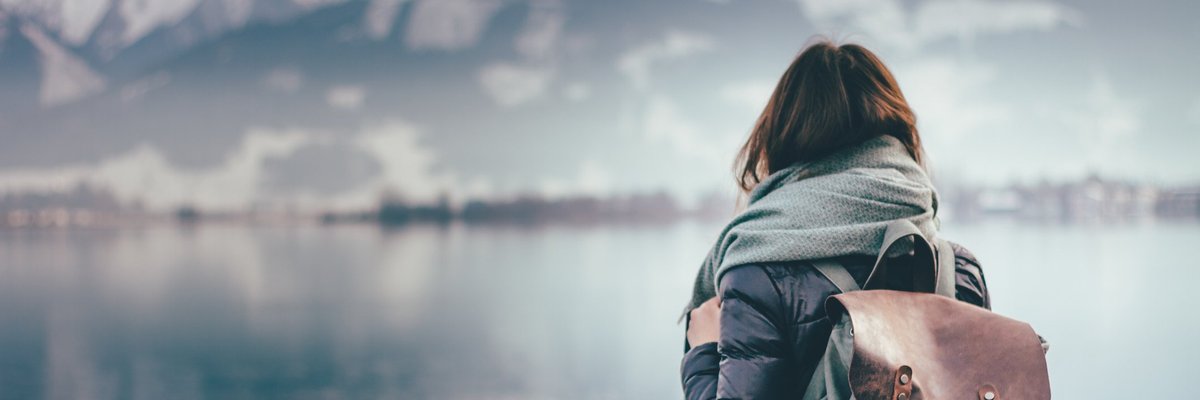 Intrepid woman wearing backpack facing away toward mountain lake.