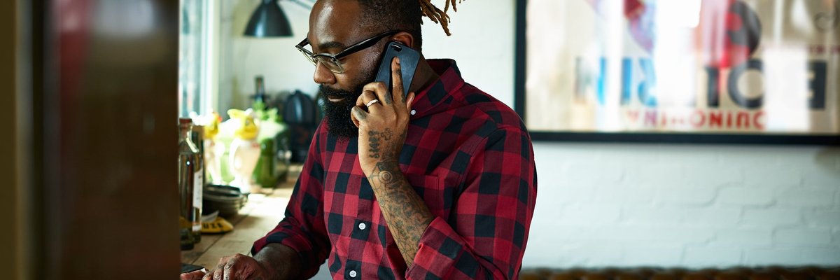 A person with a beard and tattoos talks on a smart phone while using a laptop in a design studio.