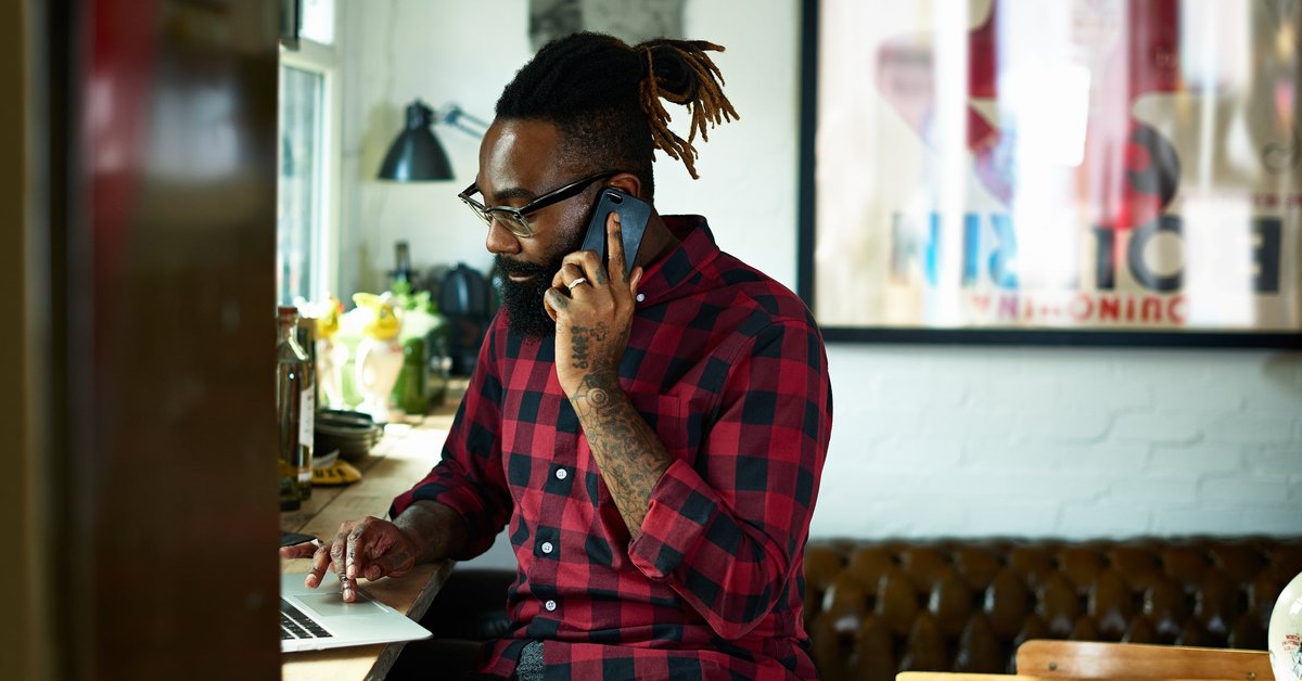 A person with a beard and tattoos talks on a smart phone while using a laptop in a design studio.