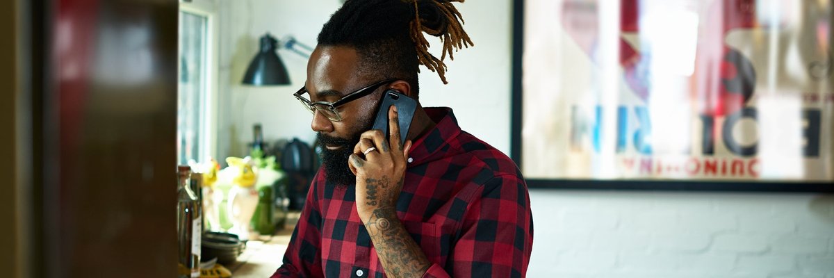 A person with a beard and tattoos talks on a smart phone while using a laptop in a design studio.