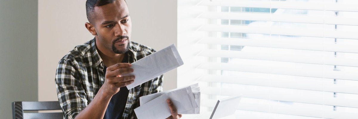 A person with facial hair sorts through bills at the kitchen table with a neutral expression.