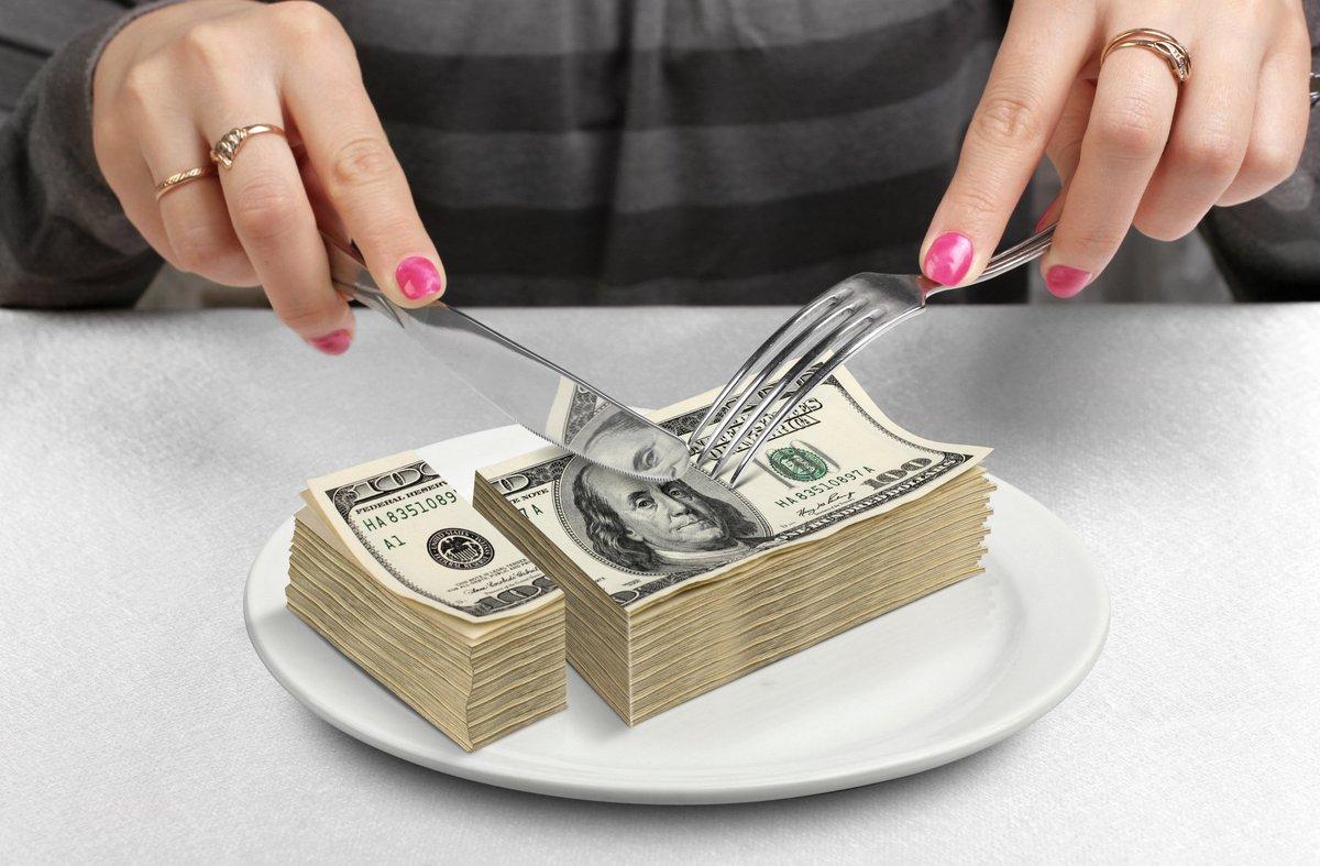 Pair of nail-polished hands cutting into a stack of 100-dollar bills with a fork and knife.
