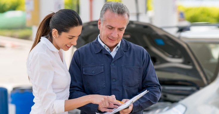Young woman at a garage talking to a mechanic about her car. They both point at paperwork.
