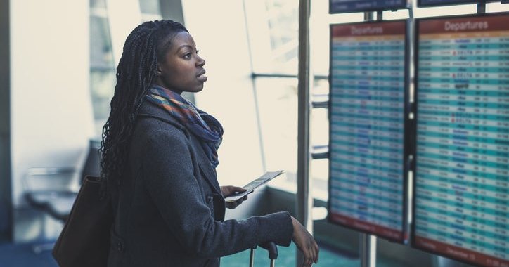 A woman at the airport checks the arrivals and departures board, holding her plane ticket and suitcase.