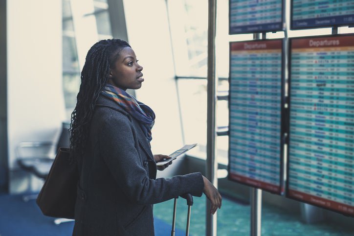 A woman at the airport checks the arrivals and departures board, holding her plane ticket and suitcase.