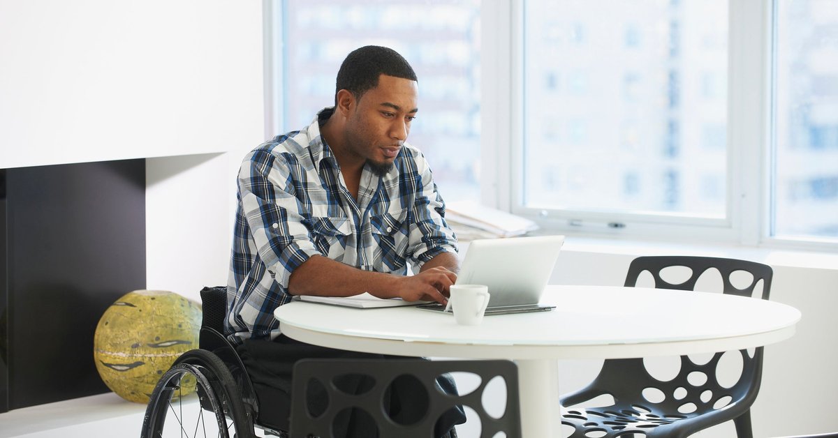 A person using a wheelchair types on a tablet while sitting at a table in a public space.