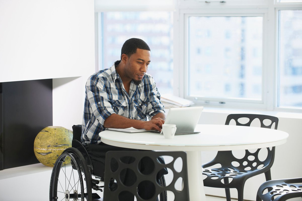 A person in a wheelchair types on a tablet while sitting at a table in a public space.