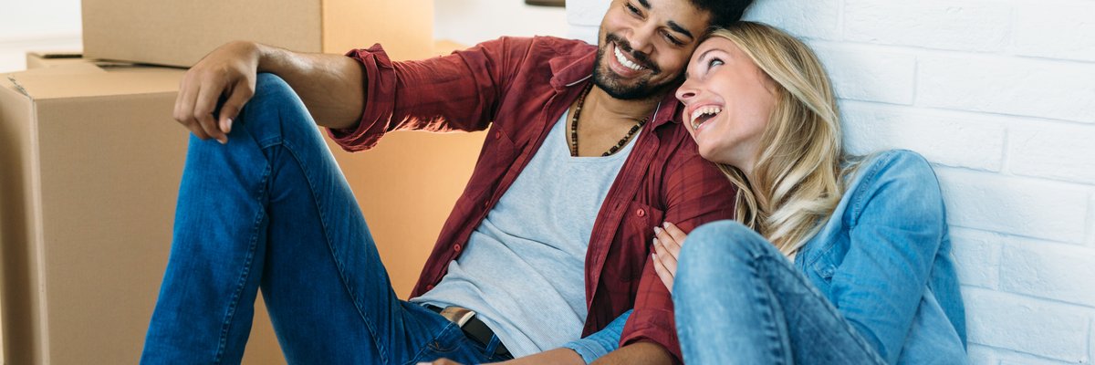 A young couple holds hands and smiles as they sit on the floor near moving boxes.
