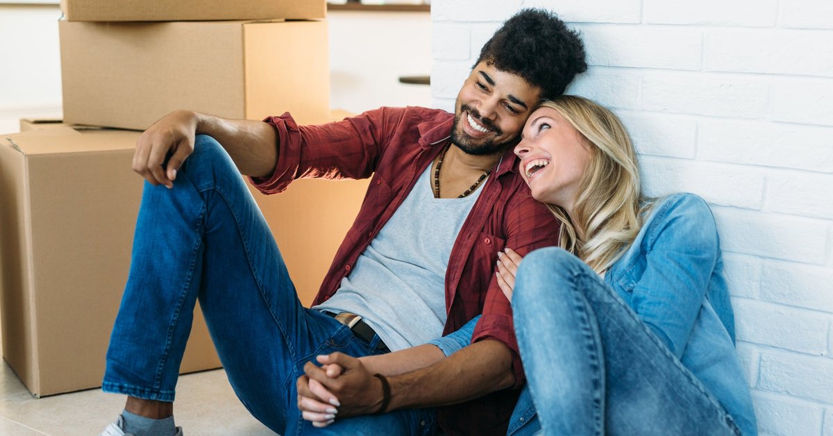 A young couple holds hands and smiles as they sit on the floor near moving boxes.