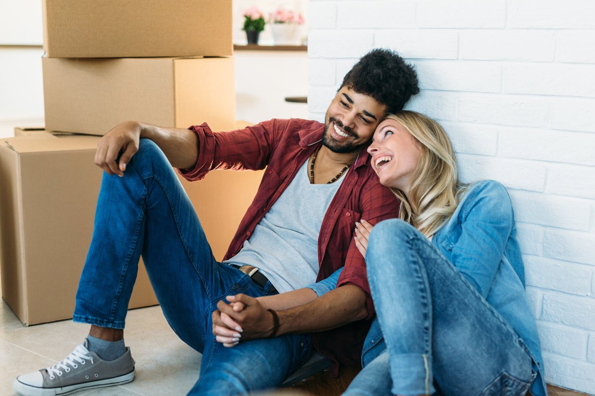 Young couple holding hands and smiling while sitting on the floor near moving boxes.
