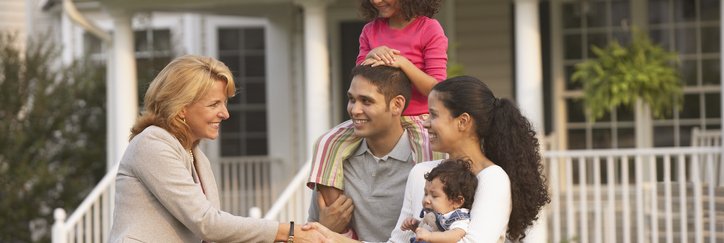 A family with children shakes hands with a real estate agent in front of a house.