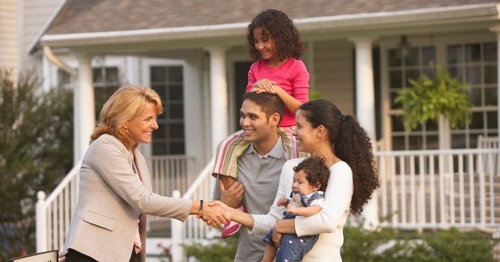 A family with children shakes hands with a real estate agent in front of a house.