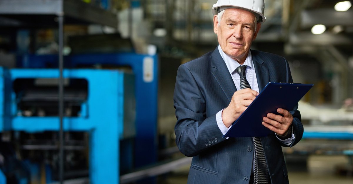 Older man in a suit and hardhat holding a clipboard.