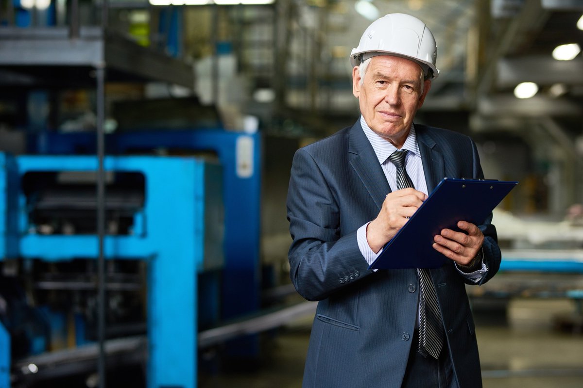 Older man in a suit and hardhat holding a clipboard.