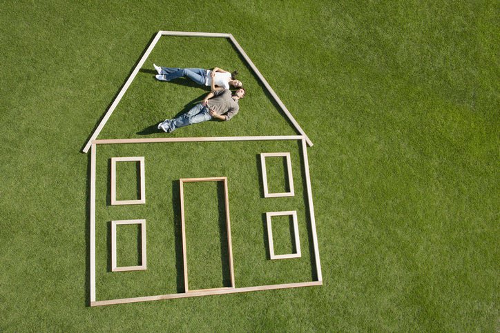 There is a couple lying on grass. An outline of a house in wood surrounds them.