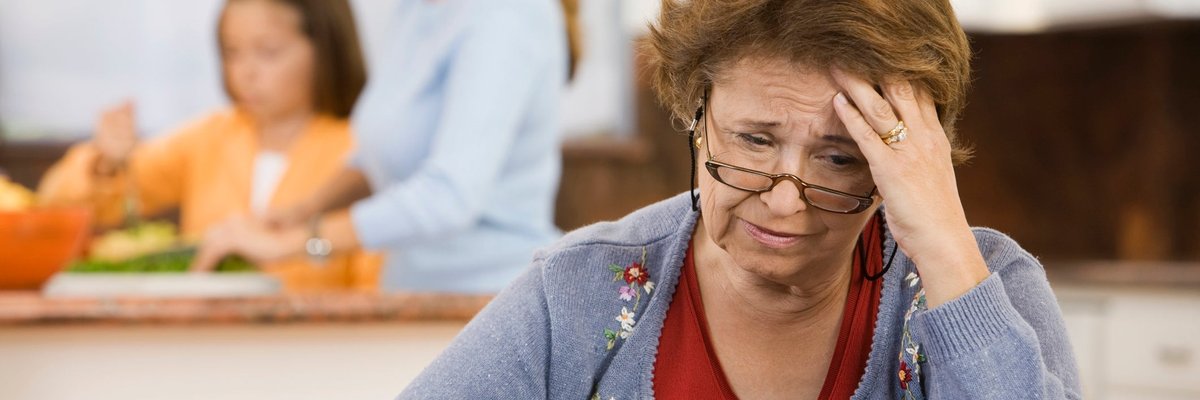 An older person worriedly reviews their checkbook in the kitchen while their family cooks in the background.
