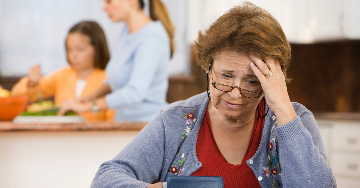 An older person worriedly reviews their checkbook in the kitchen while their family cooks in the background.